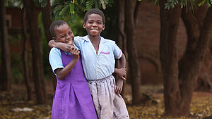 Zwei Schulkinder in Malawi lachen und umarmen sich in einer Umgebung voller Bäume und Blüten. Zwei lachende Schulkinder in Schuluniform umarmen sich unter einem Baum in Malawi und strahlen Freude und Freundschaft aus.