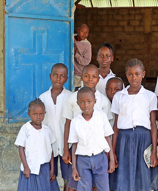 Mädchen und Jungen in Schuluniform stehen am Eingang eines Saals, an dessen Tür ein chrstliches Kreuz zu erkennen ist