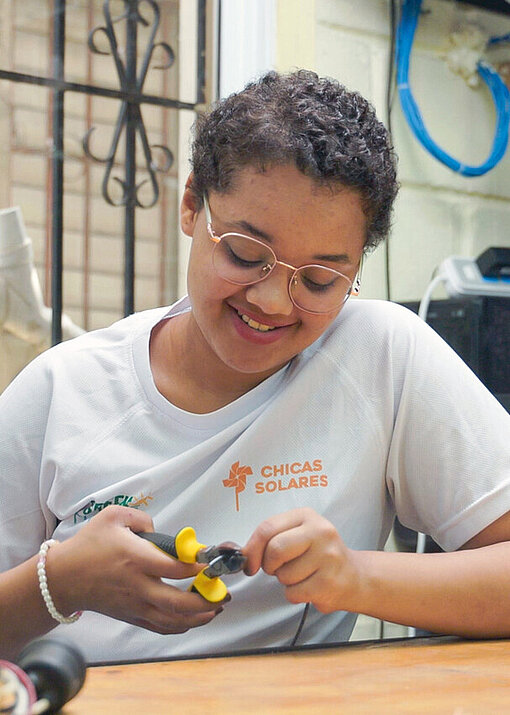 Eine junge Frau sitzt an einem Arbeitstisch und bearbeitet mit einer Zange ein Kabel; sie trägt ein Projekt‑T‑Shirt und konzentriert sich auf die praktische Übung.