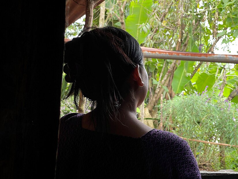 Eine junge Person steht in einem dunklen Raum und blickt durch ein Fenster nach draußen. Im Hintergrund ist üppige Vegetation mit Bananenpflanzen zu sehen. Das Gesicht ist nicht sichtbar.