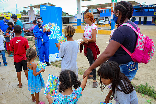 Plan Mitarbeiter:innen verteilen kindergerechtes Informationsmaterial, um geflüchtete Menschen bei ihrer Ankunft in Peru zu unterstützen. Eine Gruppe Kinder mit einer erwachsenen Begleitung bekommt von einer Plan Mitarbeiterin Comic-Hefte.