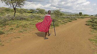 Ein schwarzer Mann in roten Gewändern und Sandalen läuft mit einem Wanderstock über einen sandigen Weg und dreht sich lächelnd zurück zur Kamera.