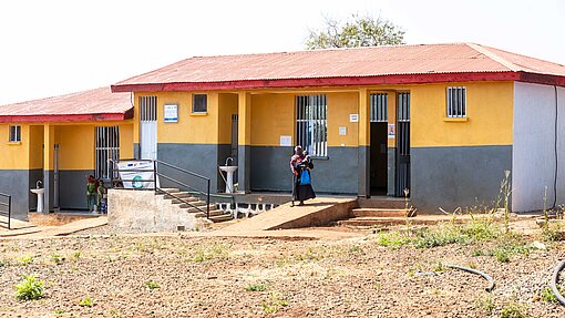 Eine Frau steht vor einem neuen Gebäude in Ura. Es ist in leuchtender Farbe gestrichen und der Boden ist noch karg von den Baunmaßnahmen.