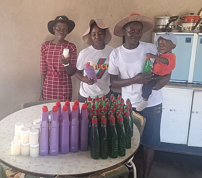 Eine Gruppe von drei Erwachsenen steht zusammen und hält Flaschen und Behälter mit Waschmittel. Sie tragen T-Shirts und Hüte und posieren für das Foto in einem Raum mit einem Tisch im Hintergrund.