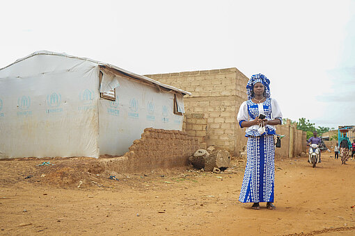 Rihanata steht auf einer Straße neben Häusern. Sie trägt ein blau-weißes Kleid und Kopftuch