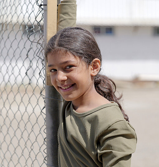 Ein Mädchen lehnt an einem Zaun aus Metall und blickt lächelnd zur Seite. Im Hintergrund ist ein helles Gebäude zu sehen.