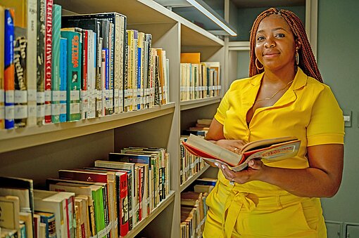 Eine dunkelhäutige Frau in leuchtend gelbem Kostüm steht an einem Bücherregal in einer Bibliothek.