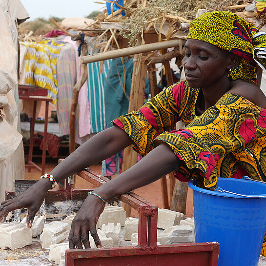 Hamssetou-schneidet-seife-202211-MLI-29-lpr.jpg Eine Frau (Hamssetou) steht an einer Werkbank und breitet Seifenstücke aus, um sie in verkaufsfertige Portionen zu zerschneiden.
