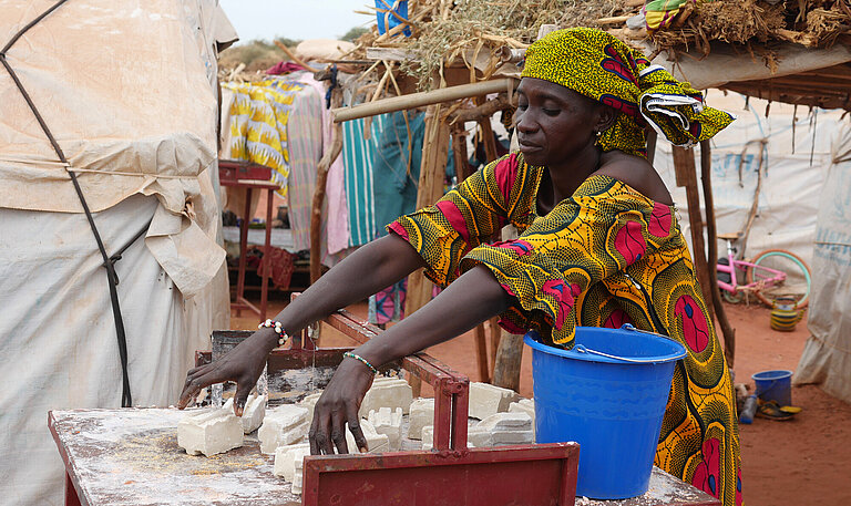 Eine Frau (Hamssetou) steht an einer Werkbank und breitet Seifenstücke aus, um sie in verkaufsfertige Portionen zu zerschneiden.