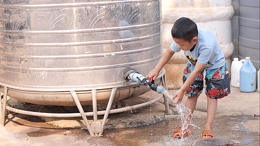 Dank der neuen Wasserstellen haben Familien kürzere Wege und besseren Zugang zu Trinkwasser. Ein Junge steht vor einem großen Wasserbehälter und wäscht sich die Hände.