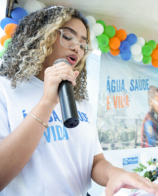 Sanmya spricht auf einer Veranstaltung des Plan-Projekts für Wasserversorgung. Eine junge Frau (Sanmya) spricht in ein Mikrofon. Im Hintergrund hägt ein Plakat und Luftballons.