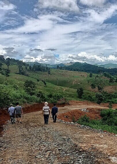 Ein Projektteam von Plan International auf dem Weg in das Dorf Huaymong, Provinz Bokeo Einige Männer gehen einen Schotterweg bergab. Er befindet sich inmitten von begrünten Hügeln.