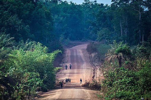 Früher Kampfgebiet, heute Handelsweg: Eine Fernstraße im westafrikanischen Liberia Die Kamera zoomt auf eine rot-braue Sandstraße, auf der Personen laufen, umgeben von grünen Bäumen