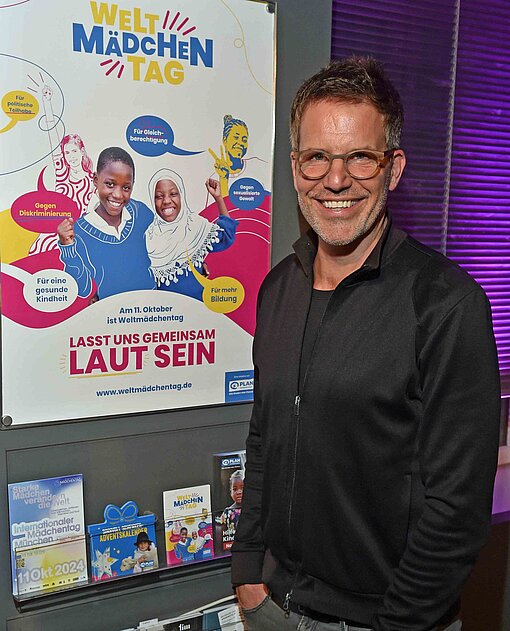 Schauspieler Tim Bergmann steht mit den Händen in der Hosentasche lächelnd vor einem Plakat zum Weltmädchentag