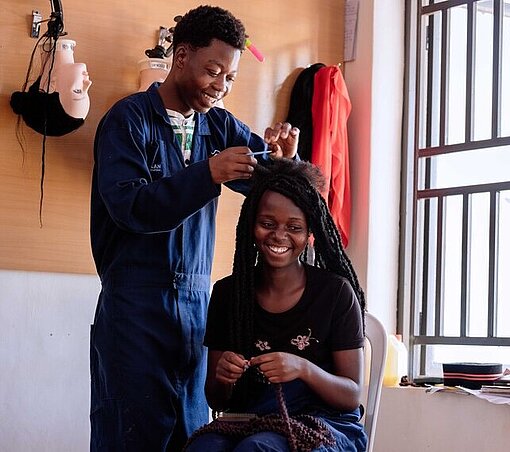 Ein junger Mann flechtet die Haare einer jungen Frau, die vor ihm auf einem Stuhl sitzt