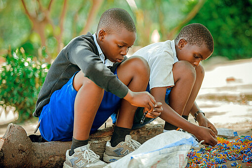 Zwei Jungen aus Tansania sitzen auf einem Balken und basteln Freundschaftsbändchen