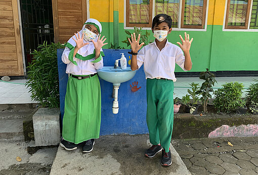 Nadia und Ceni vor dem Waschbecken der Schule Nadia und Ceni stehen neben einem Waschbecken und winken in die Kamera. Sie tragen medizinische Masken und ihre Schuluniform.