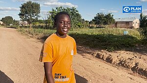 Lucy aus Sambia lächelt in die Kamera. Sie trägt ein oranges Shirt und läuft draußen.