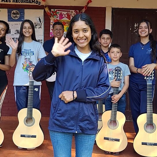 Eine Gruppe Jugendlicher steht draußen, vier von ihnen haben Gitarren vor sich stehen, ein Mädchen steht im Vordergrund und winkt in die Kamera