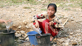 Ein Mädchen sitzt in einem ausgetrockneten Bach auf dem Boden und füllt Wasser in Plastikeimer