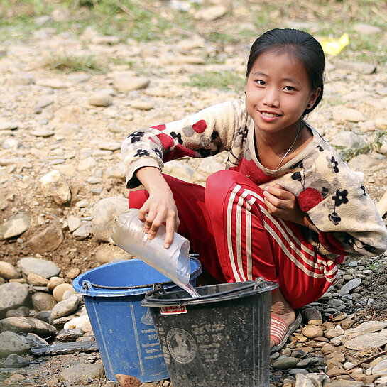 Ein Mädchen sitzt in einem ausgetrockneten Bach auf dem Boden und füllt Wasser in Plastikeimer