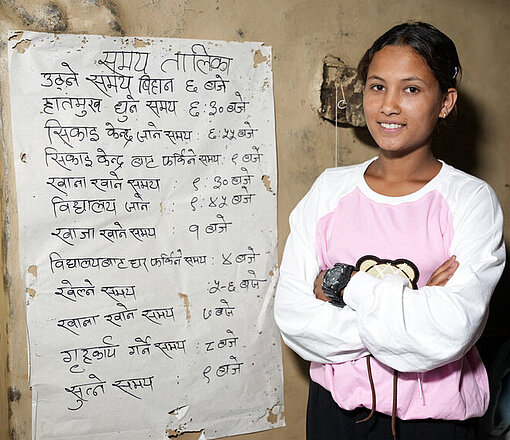 Pabitra steht in ihrer Leseecke vor einem selbst gestalteten Plakat mit handschriftlich notierten Lerninhalten und blickt in die Kamera.