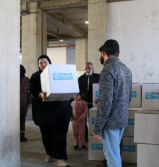 Eine libanesische Frau trägt eine Kiste mit Hilfsgütern von Plan International