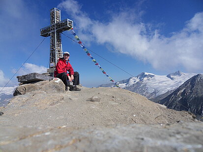 Ein Mann, Andream, sitzt an einem Gipfelkreuz und lächelt in die Kamera. Im Hintergrund sind weitere Berge zu sehen.