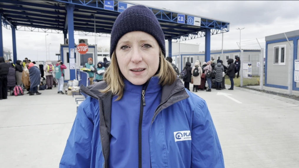 Eine Frau mit Mütze und blauer Plan-Jacke steht vor einem Grenzübergang