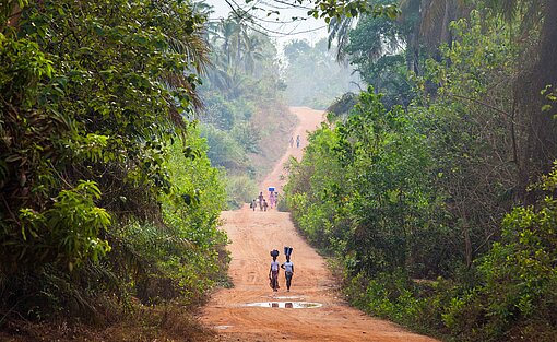 Eine Landstraße im ländlichen Guinea Mitten durch dichten Wald führt eine Straße aus rotem Lehm, auf der Personen laufen