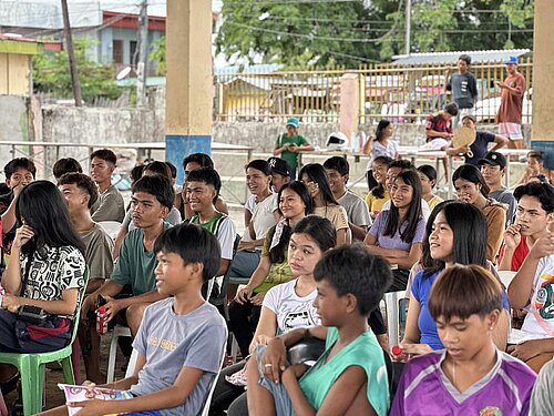Eine Gruppe von etwa 30 Schüler:innen sitzt auf Stühlen in einem Klassenzimmer. Sie hören aufmerksam zu, einige haben Hefte oder Bücher vor sich.