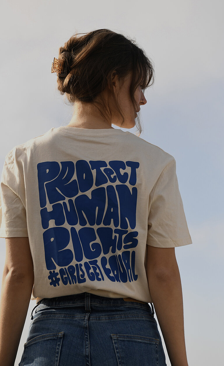 Hannah steht mit dem Rücken zur Kamera. Ihr T-Shirt ist blau bedruckt mit der Aufschrift "Protect Human Rights #Girlsgetequal"