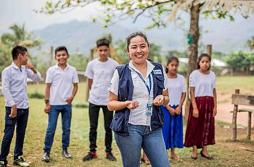 Von den Plan-Projekten profitieren wie hier in Guatemala immer gleichberechtigt Mädchen und Jungen Eine Frau mit einer blauen Weste, auf der das Logo von Plan International eingestickt ist, lächelt zusammen mit einer Gruppe Kinder