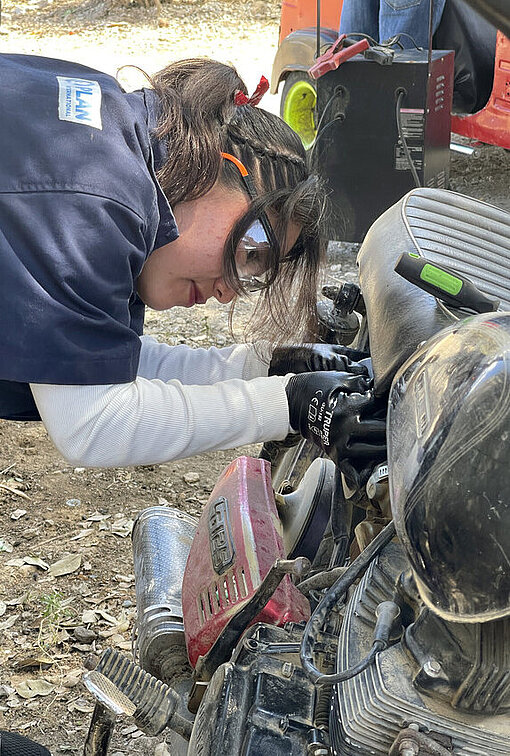 Eine junge Frau schraubt mit Schutzbrille und Arbeitskleidung ausgestattet an einem Motorrad