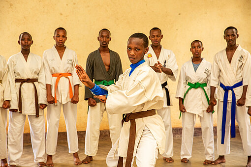Eine junge Frau macht Karate. Hinter ihr steht eine Gruppe Jugendlicher, ebenfalls im Anzug, und schaut ihr zu.