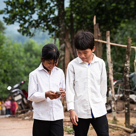 Ein Junge und ein Mädchen schauen gemeinsam auf ein Handy. Sie lächeln und tragen weiße Hemden.