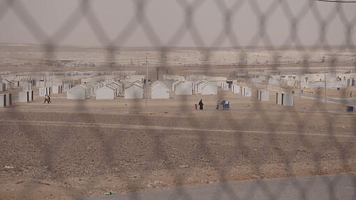 Durch einen Maschendrahtzaun hindurch sieht man Reihenweise weiße Blech-Häuser, die in der Wüste stehen.