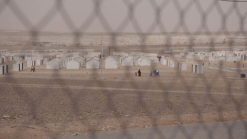 Das Azraq-Camp liegt mitten in der jordanischen Wüste Durch einen Maschendrahtzaun hindurch sieht man Reihenweise weiße Blech-Häuser, die in der Wüste stehen.