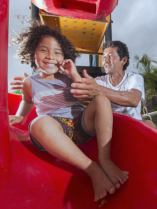 Ein Mann steht an einer roten Plastikrutsche und behütet seine rutschende Tochter mit lockigen Haaren
