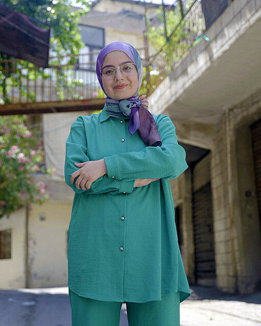 Eine junge Frau aus dem Libanon steht auf der Straße, hat die Arme vor der Brust verschränkt und schaut lächelnd in die Kamera