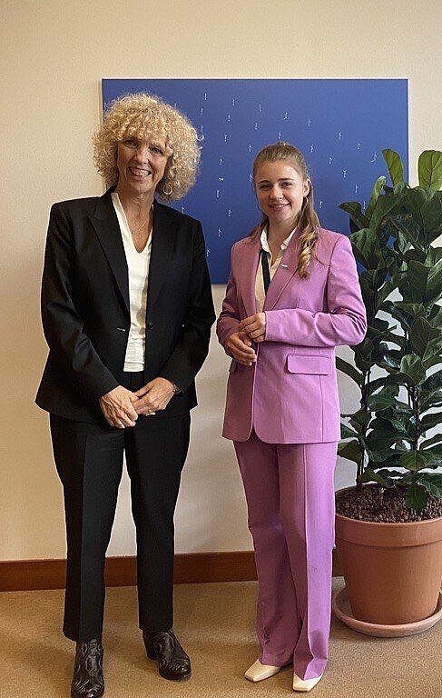 Jennifer Morgan und Kim stehen in einem Büro neben einer großen Topfpflanze. Beide tragen Anzüge und lächeln in die Kamera.