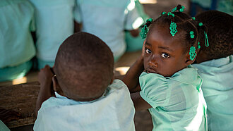 Kinder sitzen an einer Schulbank, ein kleines Mädchen guckt ernst in die Kamera.