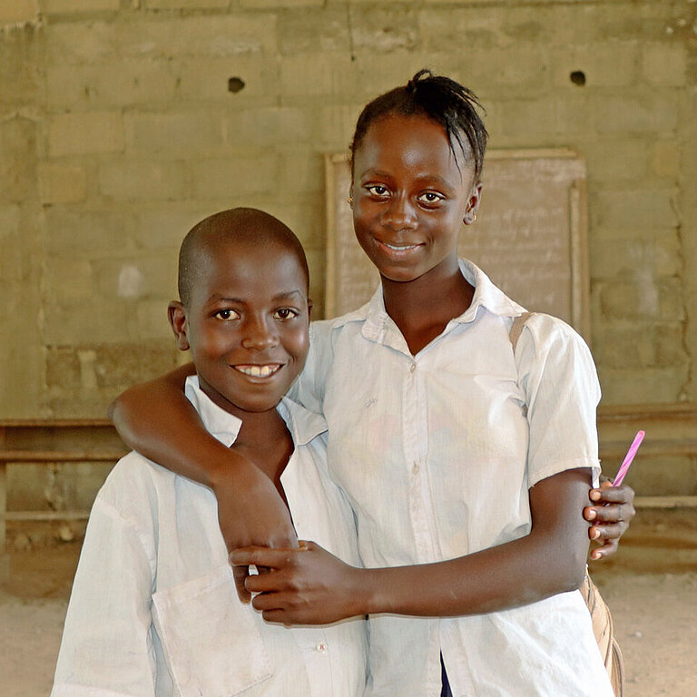 In einem kahlen Raum stehen ein Mädchen und ein Junge vor einer improvisierten Schultafel