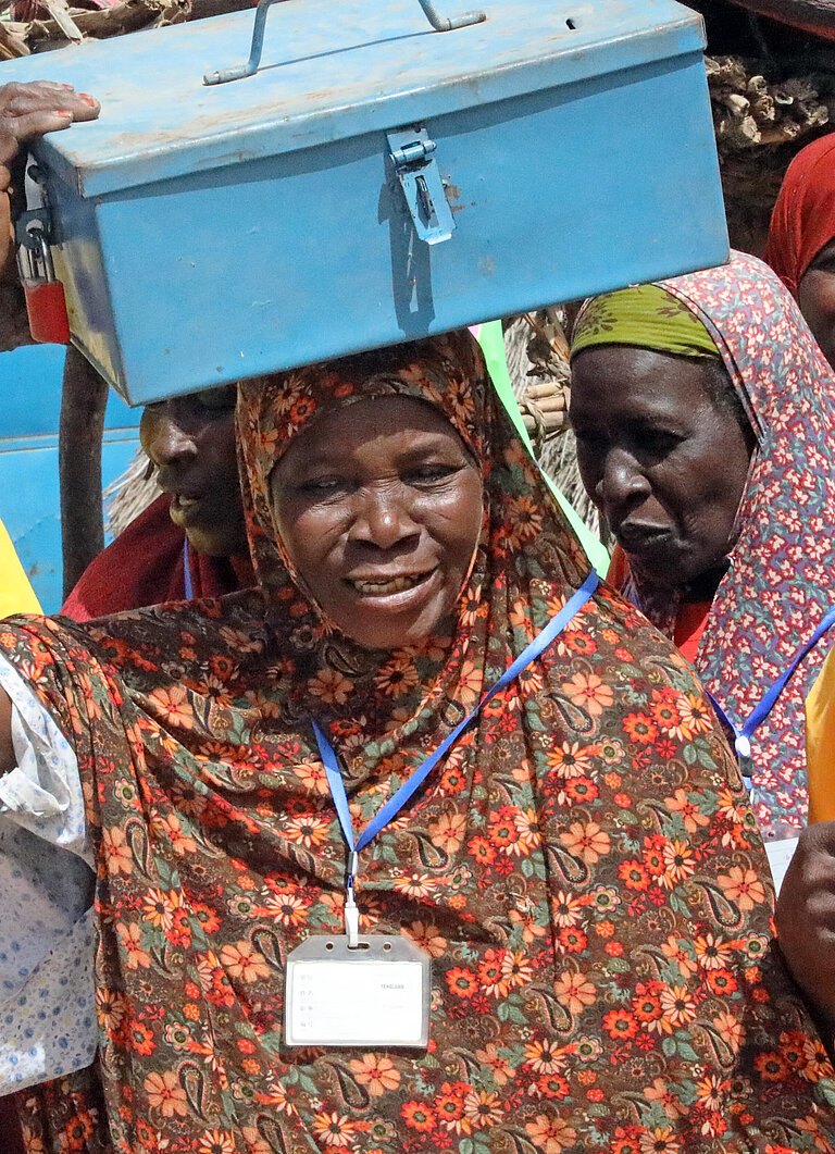 Eine Gruppe von Frauen in bunten Gewändern hat sich auf einem Dorfplatz versammelt. Eine Frau in der Mitte der Gruppe trägt eine metallene Geldkassette auf dem Kopf.