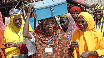 Eine Gruppe von Frauen in bunten Gewändern hat sich auf einem Dorfplatz versammelt. Eine Frau in der Mitte der Gruppe trägt eine metallene Geldkassette auf dem Kopf.