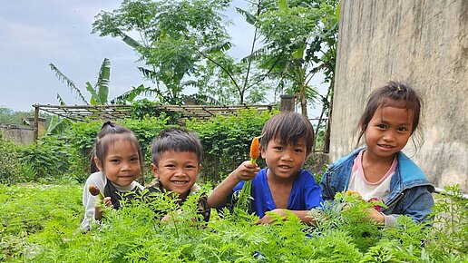Vier Kinder aus Vietnam sitzen in einem Beet und ein Kind hält stolz eine Karotte in der Hand, die es geerntet hat.