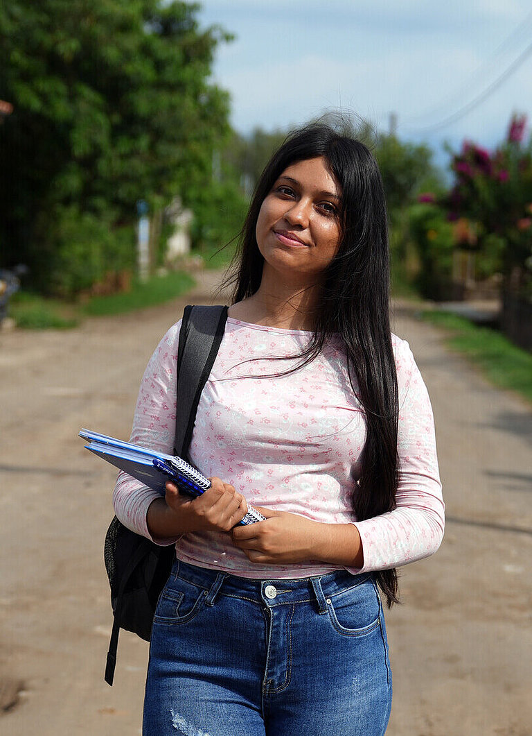Teenagerin trägt Schulrucksack und Notizblock, unterwegs in einer ländlichen Gegend. Bildung als Schlüssel für die Zukunft.
