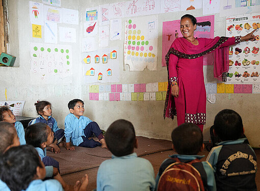Lehrerin Muna steht vor einer Schulklasse und erklärt jungen Schülerinnen und Schülern anhand von Abbildungen verschiedene Obstsorten