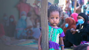 Bild: Ein kleines Kind guckt ernst in die Kamera, im Hintergrund sind andere Personen erkennbar.