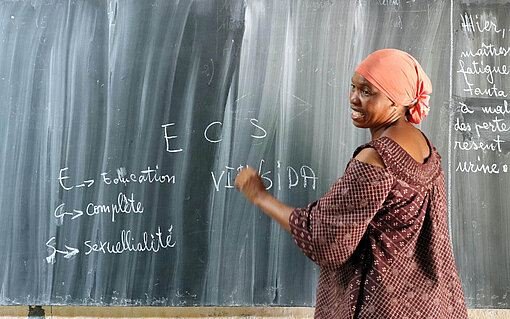 Grundschullehrerin Fanta sorgt im Unterricht für eine umfassende Sexualerziehung (Education Complète Sexuellialité, ECS) Eine Frau in rot-braunem Kleid schreibt an einer Schultafel